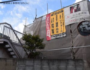 足場と飛散防止シートで覆われた施工中の建物。八陣建設の広告幕が掛かっている